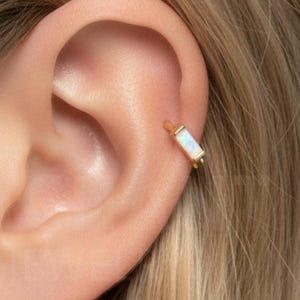 Könnte beinhalten: Ein goldfarbener Ohrklemme mit einem rechteckigen, opalisierenden Stein ist am Ohr einer Person mit blondem Haar zu sehen. Der Ohrschmuck ist ein schlichtes, elegantes Design, perfekt, um einen Hauch von Glanz zu verleihen.