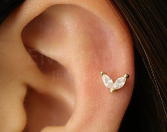 May include: A close-up of a person's ear with a gold and clear marquise shaped gemstone stud earring. The earring is in the shape of a V.