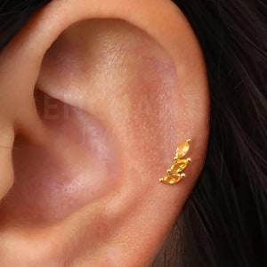 Può includere: Un piercing all'orecchio dorato con tre pietre preziose color ambra, taglio marquise, disposte in un design curvo a forma di foglia. Il gioiello è posto sulla pelle di un orecchio.