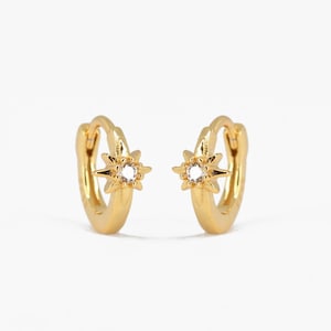 Peut inclure: Une paire de boucles d'oreilles créoles dorées avec un petit charme en forme d'étoile orné d'un strass transparent au centre.