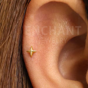 Könnte beinhalten: Nahaufnahme eines Ohrs mit einem kleinen, goldfarbenen sternförmigen Ohrring. Der Ohrring ist ein vierzackiger Stern. Der Text "ENCHANT JEWELRY" ist im Hintergrund sichtbar.