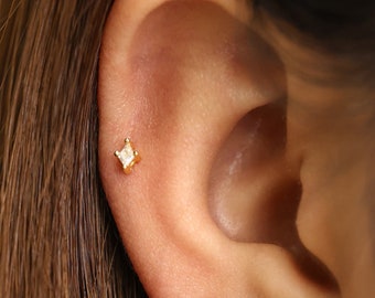 May include: A close-up of a person's ear with a small, square-shaped diamond earring in their earlobe.