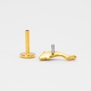 May include: Gold dolphin body jewelry with a screw-on post. The dolphin is facing left and has a textured surface.