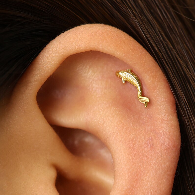 May include: Gold fish shaped cartilage earring. The earring is a small, detailed fish with a textured surface.