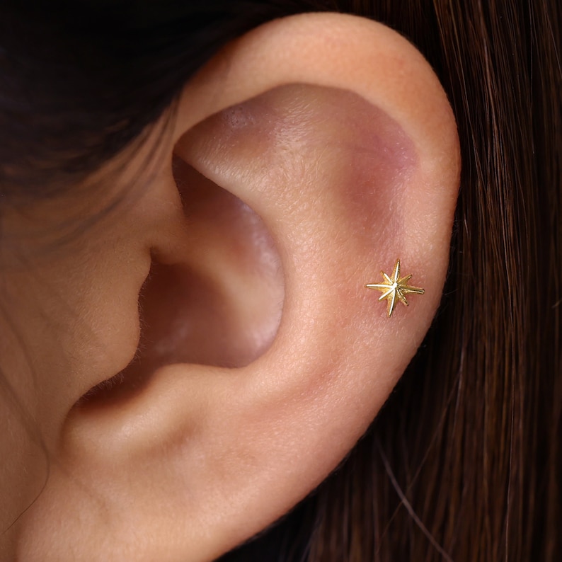 Pode incluir: Um close-up de um brinco dourado em forma de estrela. O brinco &eacute; pequeno e tem v&aacute;rias pontas, dando-lhe uma apar&ecirc;ncia de explos&atilde;o estelar. O brinco &eacute; colocado na orelha, destacando o seu design delicado e brilho met&aacute;lico.