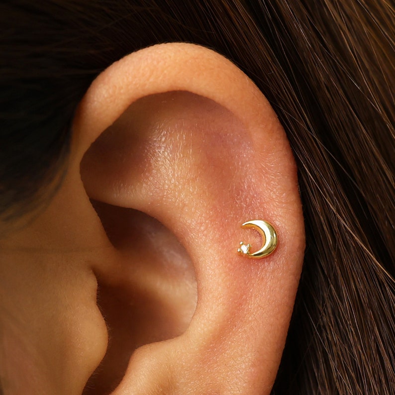 May include: A gold crescent moon earring with a small diamond accent. The earring is worn in the upper cartilage of the ear.