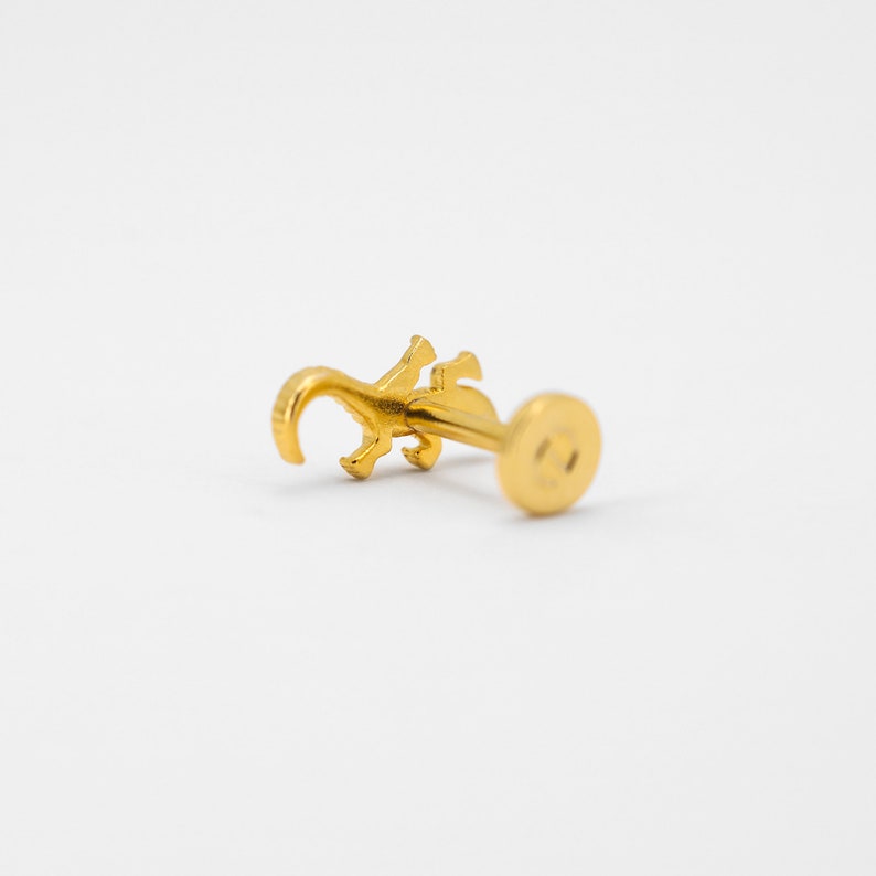 May include: Gold-toned body piercing featuring a small lizard design. The piercing is made of metal and has a round backing.