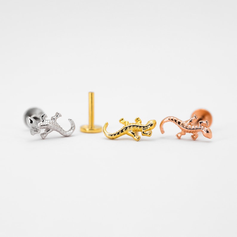 May include: Three silver, gold, and rose gold lizard body piercing jewelry pieces. Each piece features a small lizard design.