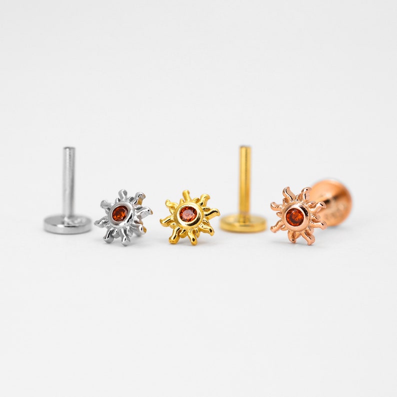 Op de afbeelding: Drie zonvormige lichaamsieraden met een rode edelsteen in het midden. De stukken zijn gemaakt van zilver, goud en ros&eacute;goud.