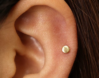 May include: A close-up of a person's ear with a small, round, gold stud earring.