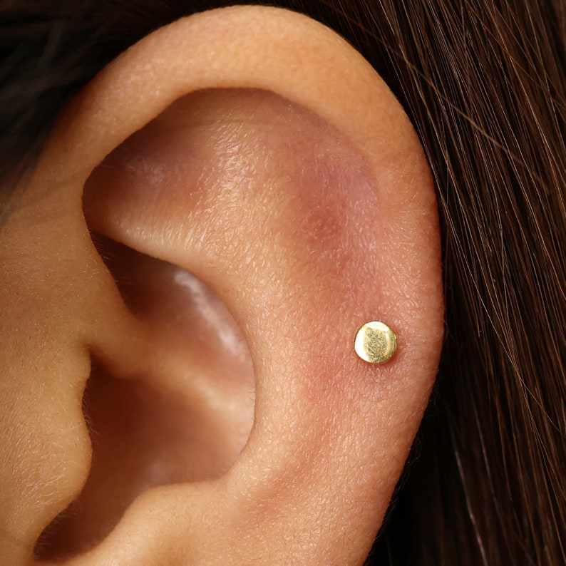 May include: A close-up of a person's ear with a small, round, gold stud earring.