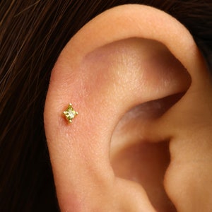 May include: A close-up of a person's ear with a small, square-shaped, yellow gemstone stud earring.