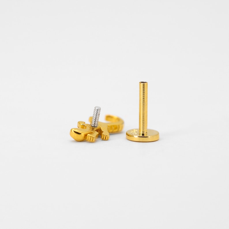 May include: Gold body jewelry with a small, detailed, gold lizard design. The jewelry is a labret stud with a flat back and a threaded post.