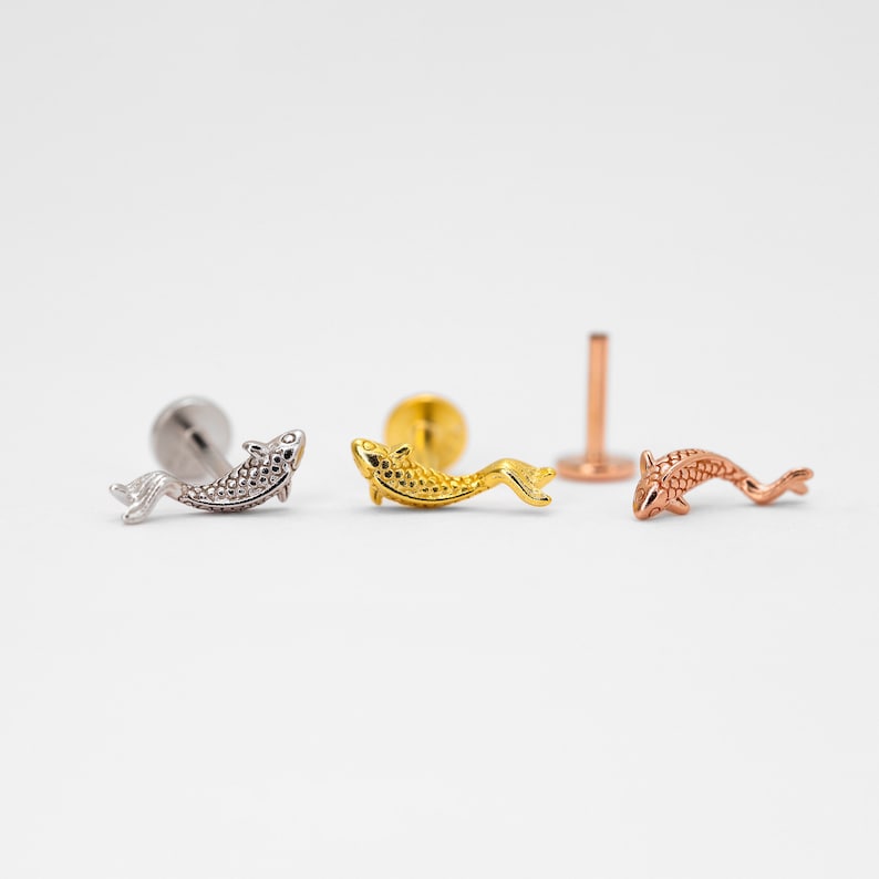 May include: Three gold fish charm body jewelry pieces. The charms are in silver, gold, and rose gold. The charms are in the shape of a fish with scales.