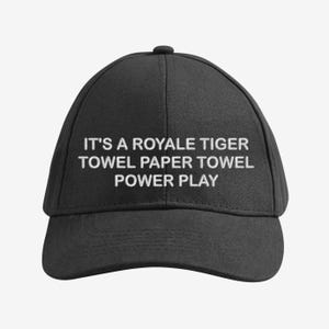 Può includere: Cappellino da baseball nero con testo bianco ricamato: "IT'S A ROYALE TIGER TOWEL PAPER TOWEL POWER PLAY". Il cappellino ha una visiera curva e un bottone sulla parte superiore.