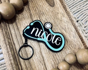 Può includere: Un portachiavi a forma di stetoscopio nero e turchese con il nome "nicole" scritto in lettere bianche.