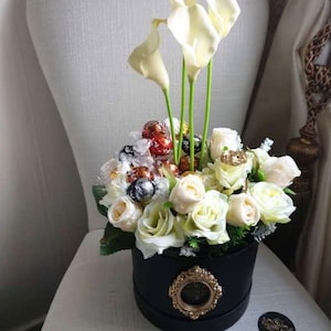 May include: A black hat box filled with white roses and white calla lilies. The box is decorated with a gold medallion and the flowers are surrounded by chocolate candies.