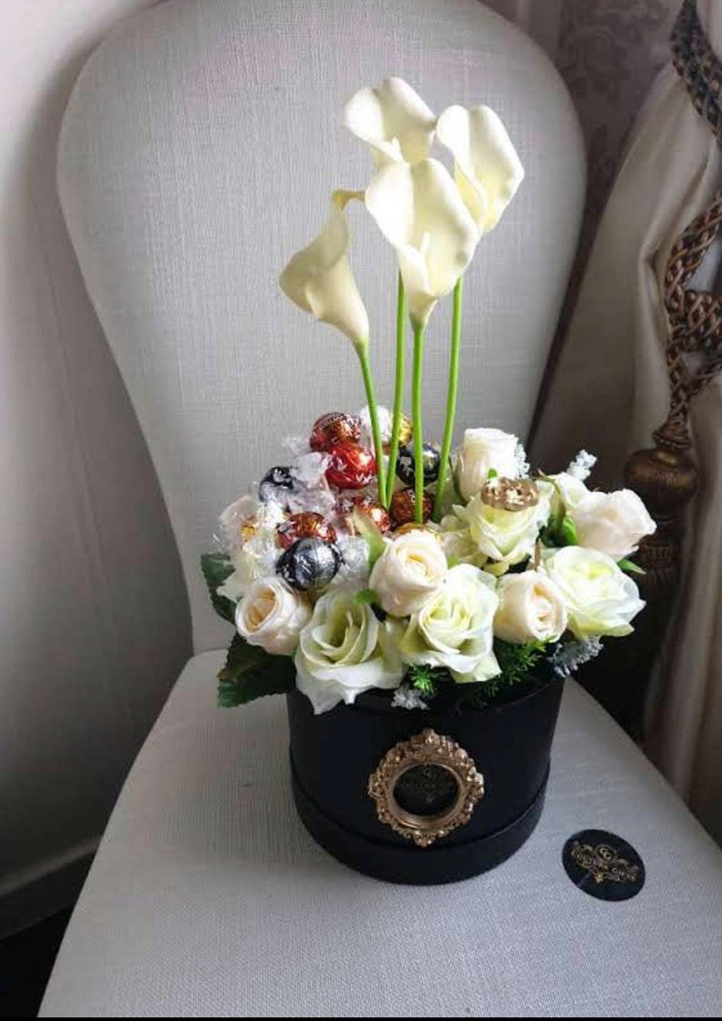 May include: A black hat box filled with white roses and white calla lilies. The box is decorated with a gold medallion and the flowers are surrounded by chocolate candies.