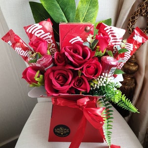 Puede incluir: Una caja roja llena de rosas rojas y chocolates Maltesers. La caja está atada con una cinta roja y tiene una pegatina que dice "Gourmet Gifts".