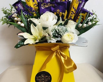 May include: A yellow box filled with white flowers and a variety of Cadbury Dairy Milk chocolate bars. The box is tied with a gold ribbon and has a black circle sticker with the text "Couture Gifts" and a website address.