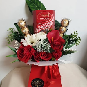 Pode incluir: Uma caixa de presente vermelha e branca cheia de rosas vermelhas, flores brancas e uma caixa de trufas de chocolate ao leite Lindt Lindor. A caixa tem uma fita vermelha e um adesivo dourado que diz "Couture Gifts".