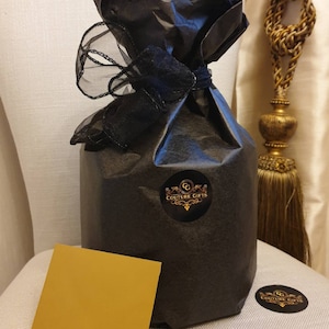 May include: A black gift bag with a black ribbon and a gold sticker that says "Couture Gifts". The bag is sitting on a white surface with a gold card.