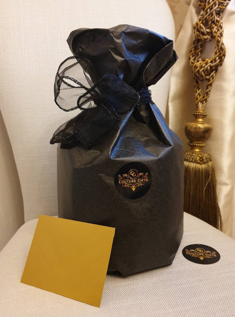 May include: A black gift bag with a black ribbon and a gold sticker that says "Couture Gifts". The bag is sitting on a white surface with a gold card.