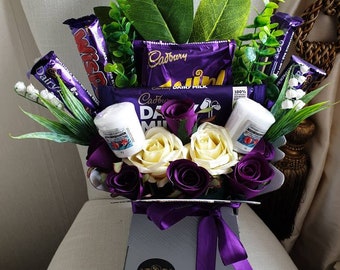 Puede incluir: Un ramo de chocolate morado y blanco con barras de chocolate Cadbury Dairy Milk, envuelto en papel de seda morado y dispuesto en una caja gris con una cinta morada. El ramo también incluye rosas blancas y moradas y hojas verdes.