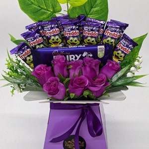 May include: A purple gift box with a purple ribbon and a bouquet of purple roses. The box is filled with Cadbury Dairy Milk chocolate bars and Freddo chocolate frogs.