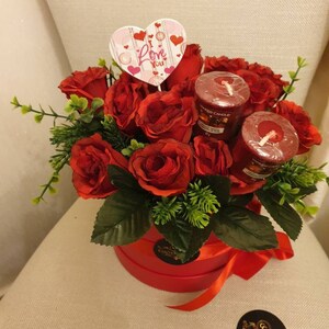 May include: A red floral arrangement in a round red box, featuring red roses, green foliage, and two red candles. A heart-shaped sign with the words "Love You" is placed in the center. A red ribbon is tied around the box.