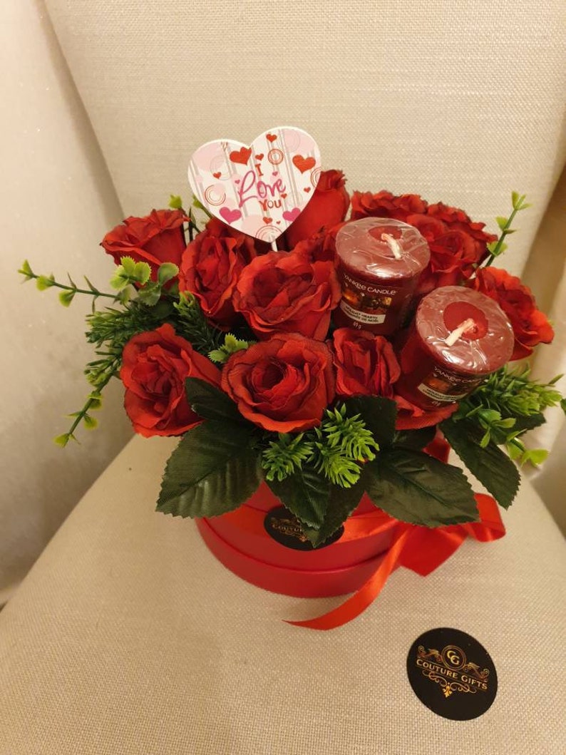 May include: A red floral arrangement in a round red box, featuring red roses, green foliage, and two red candles. A heart-shaped sign with the words "Love You" is placed in the center. A red ribbon is tied around the box.