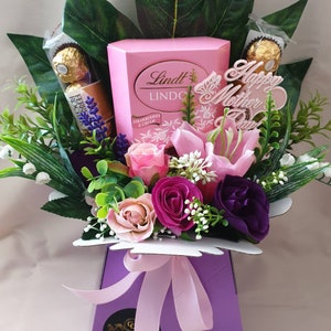 May include: A purple box with a pink ribbon and a bouquet of pink, purple, and white flowers. The box contains a box of Lindt Lindor Strawberries & Cream chocolates and a Happy Mother's Day sign.