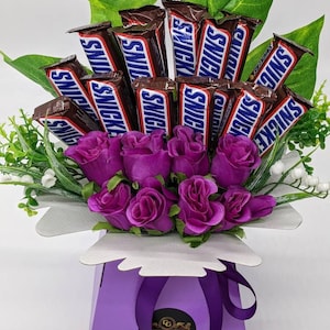 Könnte beinhalten: Eine violette Geschenkbox mit einem weißen Deckel und einem violetten Band. Die Box ist gefüllt mit einem Strauß violetter Rosen und Schokoladenriegeln, die in braune Folie verpackt sind und den Schriftzug "SNICKERS" tragen.