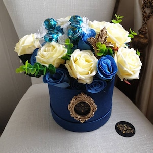May include: A blue box filled with white and blue roses and wrapped in cellophane with blue and green chocolate candies. The box has a gold emblem with the text "Couture Gift".