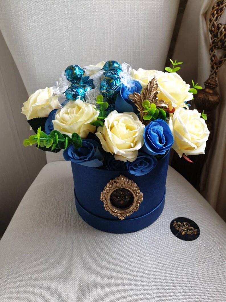 May include: A blue box filled with white and blue roses and wrapped in cellophane with blue and green chocolate candies. The box has a gold emblem with the text "Couture Gift".