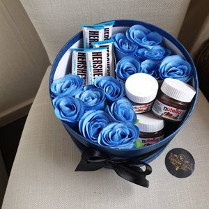 May include: A blue gift box filled with blue roses, chocolate bars, and small jars of Nutella. The box has a black ribbon and a small tag that says "Couture Gifts".