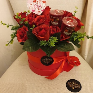 May include: A red round gift box tied with a red ribbon, filled with red roses and greenery. Includes three red Yankee Candle votives and a heart-shaped card that says "I Love You". A Couture Gifts sticker is on the box.