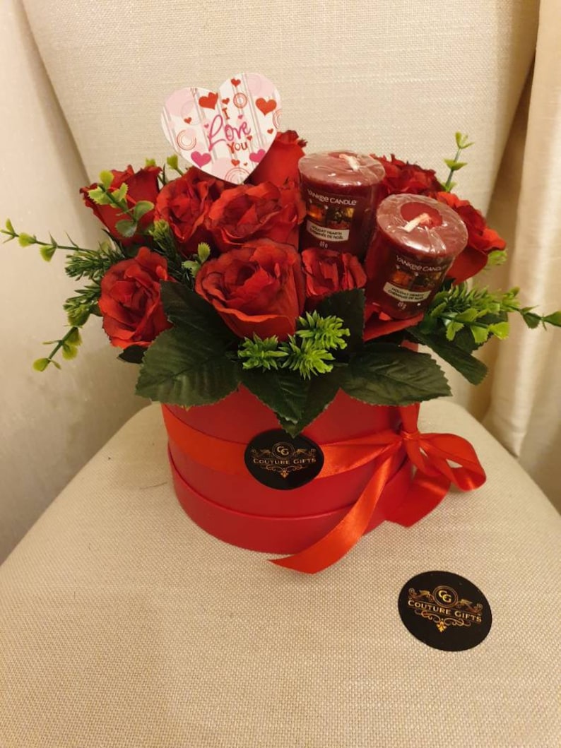 May include: A red round gift box tied with a red ribbon, filled with red roses and greenery. Includes three red Yankee Candle votives and a heart-shaped card that says "I Love You". A Couture Gifts sticker is on the box.