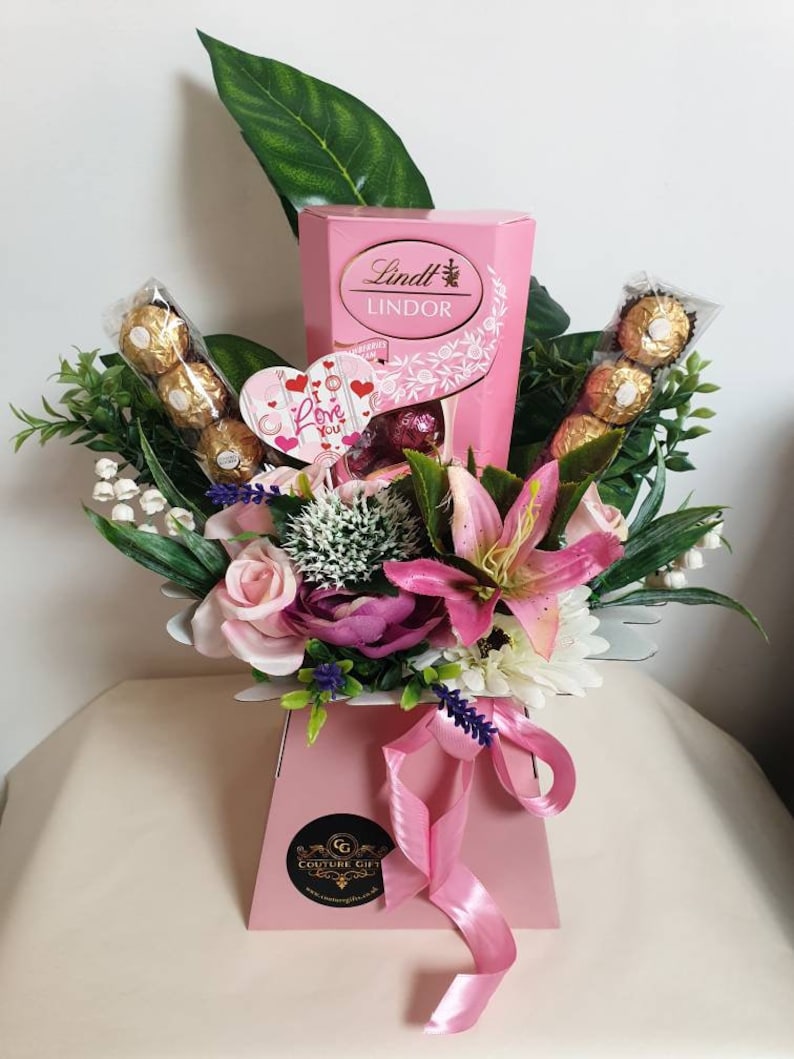 Puede incluir: Caja de regalo rosa llena de un ramo de flores rosas, blancas y moradas, una caja de chocolates Lindt Lindor y una tarjeta en forma de coraz&oacute;n que dice "Te amo".