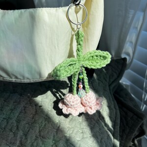 May include: A crocheted cherry keychain with green leaves and pink cherries. The keychain is attached to a silver key ring.
