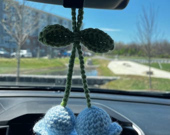 May include: A crocheted car charm with two blue bell-shaped flowers and a green bow. The charm is hanging from the rearview mirror of a car.