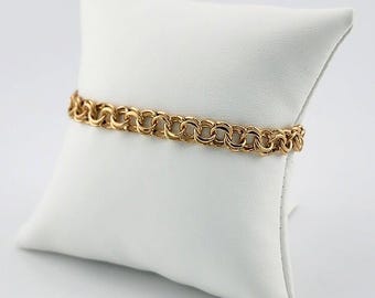 May include: A gold-colored bracelet with a double-link chain design. The bracelet is displayed on a white pillow, showcasing its intricate pattern and warm metallic hue. The links are closely connected, creating a continuous, elegant look.