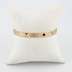 May include: A gold bracelet with a geometric pattern of alternating gold, rose gold, and silver-colored diamond shapes. The bracelet is displayed on a white pillow against a white background. The bracelet is approximately 0.25 inches wide.