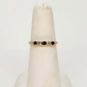May include: A gold band ring featuring three red gemstones and two clear gemstones. The ring is displayed on a white finger form. The gemstones are set in a row across the top of the ring.