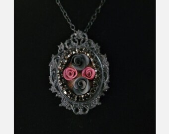 May include: A black necklace with a silver oval pendant featuring three black and pink fabric roses surrounded by black rhinestones.