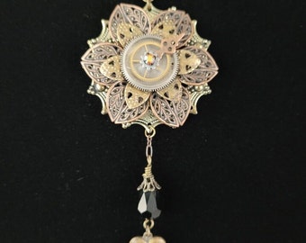 Puede incluir: Un collar de estilo steampunk en oro, plata y bronce con un colgante en forma de flor que presenta un mecanismo de relojería y una gota de cristal negro.