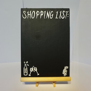Puede incluir: Pizarra negra con texto blanco que dice "SHOPPING LIST:". Hay dibujos blancos de una botella de vino, dos copas de vino y un racimo de uvas en la pizarra.