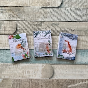 Könnte beinhalten: Drei kleine, rechteckige Deko-Boxen mit winterlichen Motiven. Die Boxen sind aus Pappe und haben einen weißen Hintergrund mit bunten Illustrationen. Die erste Box sagt "Winter" und zeigt einen Rotkehlchen. Die zweite Box sagt "Winter Wonderland" und zeigt ein Reh. Die dritte Box sagt "A Winter's Tale" und zeigt einen Fuchs.