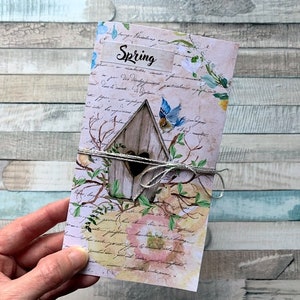 Op de afbeelding: Een roze en witte kaart met een vogelhuisje en een blauwe vogel. De kaart heeft een handgeschreven bericht met de tekst "Lente" en is vastgebonden met touw.