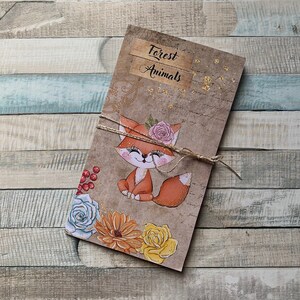 Op de afbeelding: Een bruin papieren notitieboekje met de tekst "Forest Animals" op de omslag. De omslag heeft een cartoonvos met een bloemenkrans, omringd door bloemen en bessen.